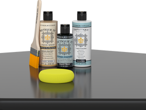 Retique It® Light Wood Wood’n Primer with Classic Black Gel Stain Alternative, gloss topcoat, brush, and yellow sponge on a reflective tabletop.