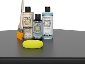 Retique It® Light Wood Wood’n Primer with Classic Black Gel Stain Alternative, satin topcoat, brush, and yellow sponge on a tabletop.