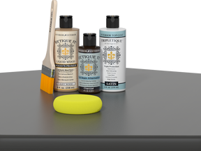 Retique It® Light Wood Wood’n Primer with Charcoal Gel Stain Alternative, satin topcoat, brush, and yellow sponge on a tabletop.