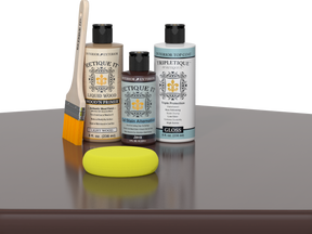 Retique It® Light Wood Wood’n Primer with Java Gel Stain Alternative, gloss topcoat, brush, and yellow sponge on a reflective tabletop.