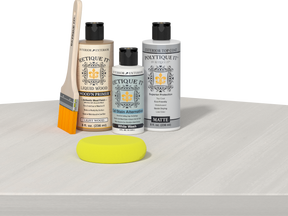 Retique It® Light Wood Wood’n Primer with White Wash Gel Stain Alternative, matte topcoat, brush, and yellow sponge on a white tabletop.