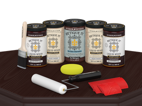 Retique It Red Mahogany Wood'n Kit featuring Red Mahogany Liquid Wood stain, Light Wood primer, Red Mahogany Gel Stain Alternative, paint brush, white roller, yellow sponge, and red graining tools displayed on a dark wood tabletop.