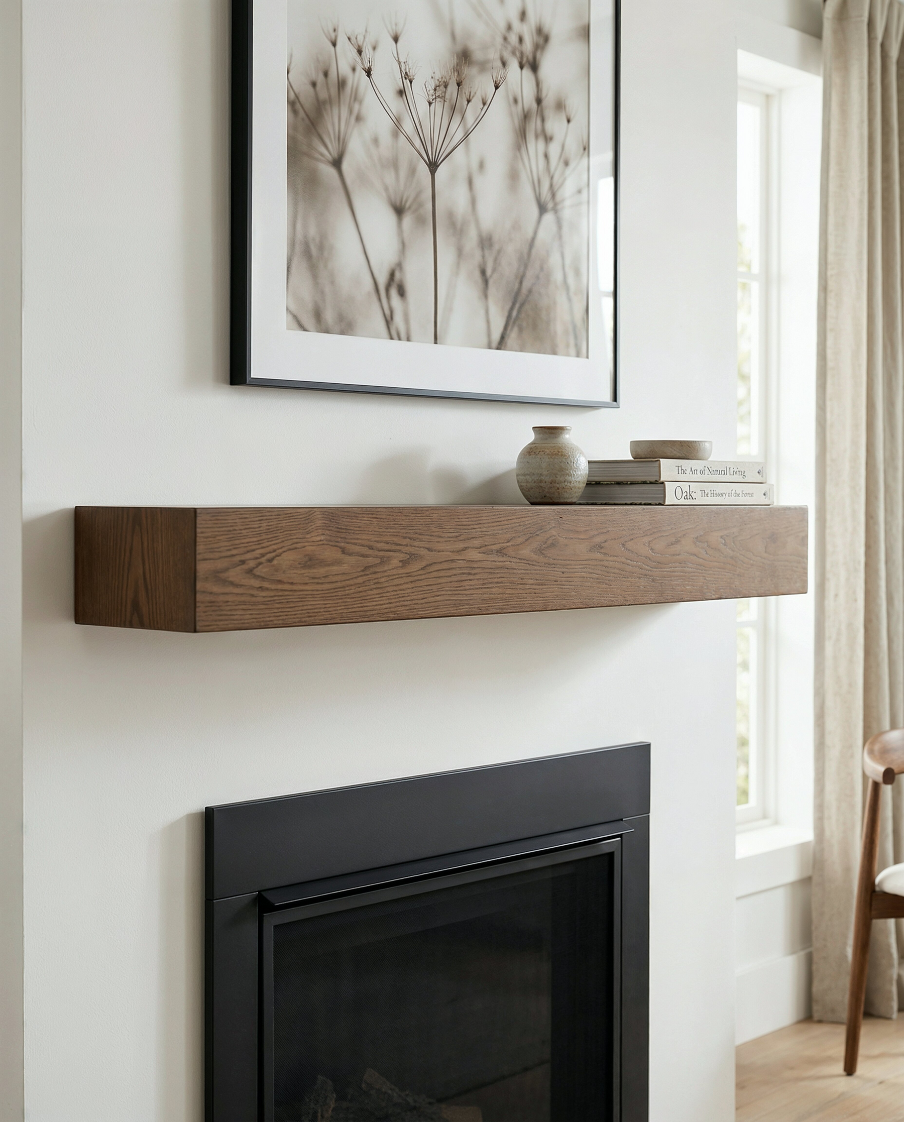 A floating wooden mantel shelf mounted on a white wall above a black fireplace, styled with a small ceramic vase and books. A framed botanical print hangs above, and a window with beige curtains is visible on the right.