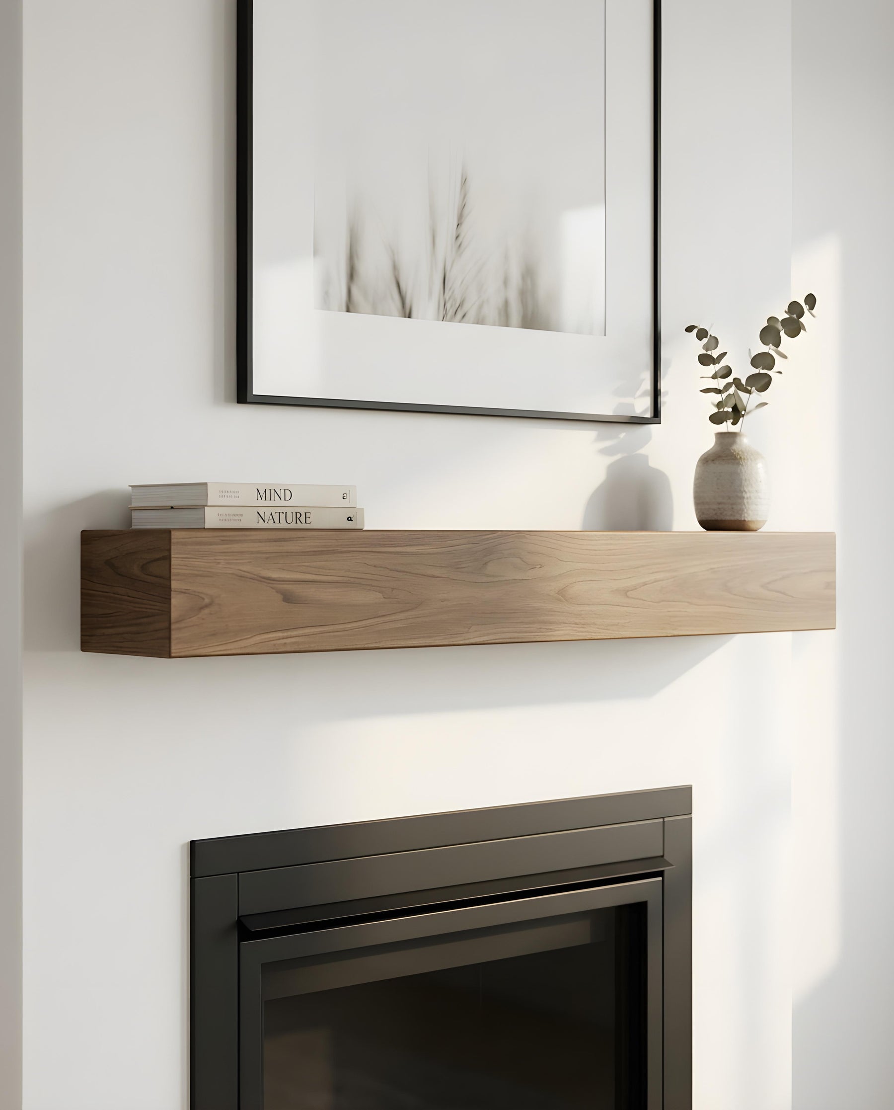 A modern floating wooden mantel shelf mounted on a white wall above a dark fireplace. The shelf holds a stack of two books and a small textured vase with dried eucalyptus stems.