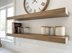 Two floating wooden kitchen shelves mounted on a white wall above a white subway tile backsplash. The top shelf is styled with a large round clock, and the lower shelf holds a cookbook and several glass canisters with wooden lids.