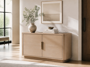 A light beige wood sideboard cabinet with two doors and vertical brass handles, styled in a bright, modern entryway with a vase of greenery, books, and a framed print on top.