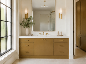 A modern double-sink bathroom vanity in a warm camel brown finish with a white countertop, set beneath a large wall mirror. The vanity features brass hardware and is styled with a vase of greenery and soap dispensers, with a large window on the left.