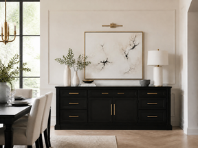 A styled interior scene showing a black sideboard or credenza with gold-tone handles, placed in a dining room beneath a large framed abstract print. The room includes a table and chairs, a lamp, vases, and a window, so this is a real environment rather than a clean studio product shot.