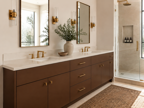 A modern double-sink bathroom vanity with dark brown cabinets, a white countertop, and gold hardware, shown in a styled bathroom setting with mirrors, sconces, and a walk-in shower.