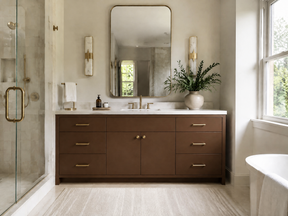 A modern bathroom vanity featuring a dark brown wood cabinet with gold handles, a white countertop, a large rectangular mirror, and a vase of greenery, set in a bright bathroom with a glass shower enclosure.