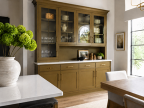 Gold-toned built-in hutch with glass-front cabinets, white countertop, and place settings in a bright dining room.
