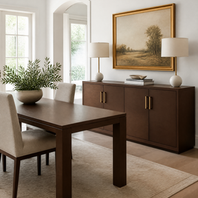 A styled dining room scene featuring a dark brown wooden dining table and matching chairs, a matching sideboard with gold handles, white table lamps, a potted green plant, and a large framed landscape painting on the wall.