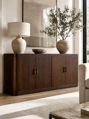 A dark brown wooden sideboard cabinet in a modern living room setting, styled with a textured ceramic table lamp, a small stone bowl, and a large vase with olive branches.