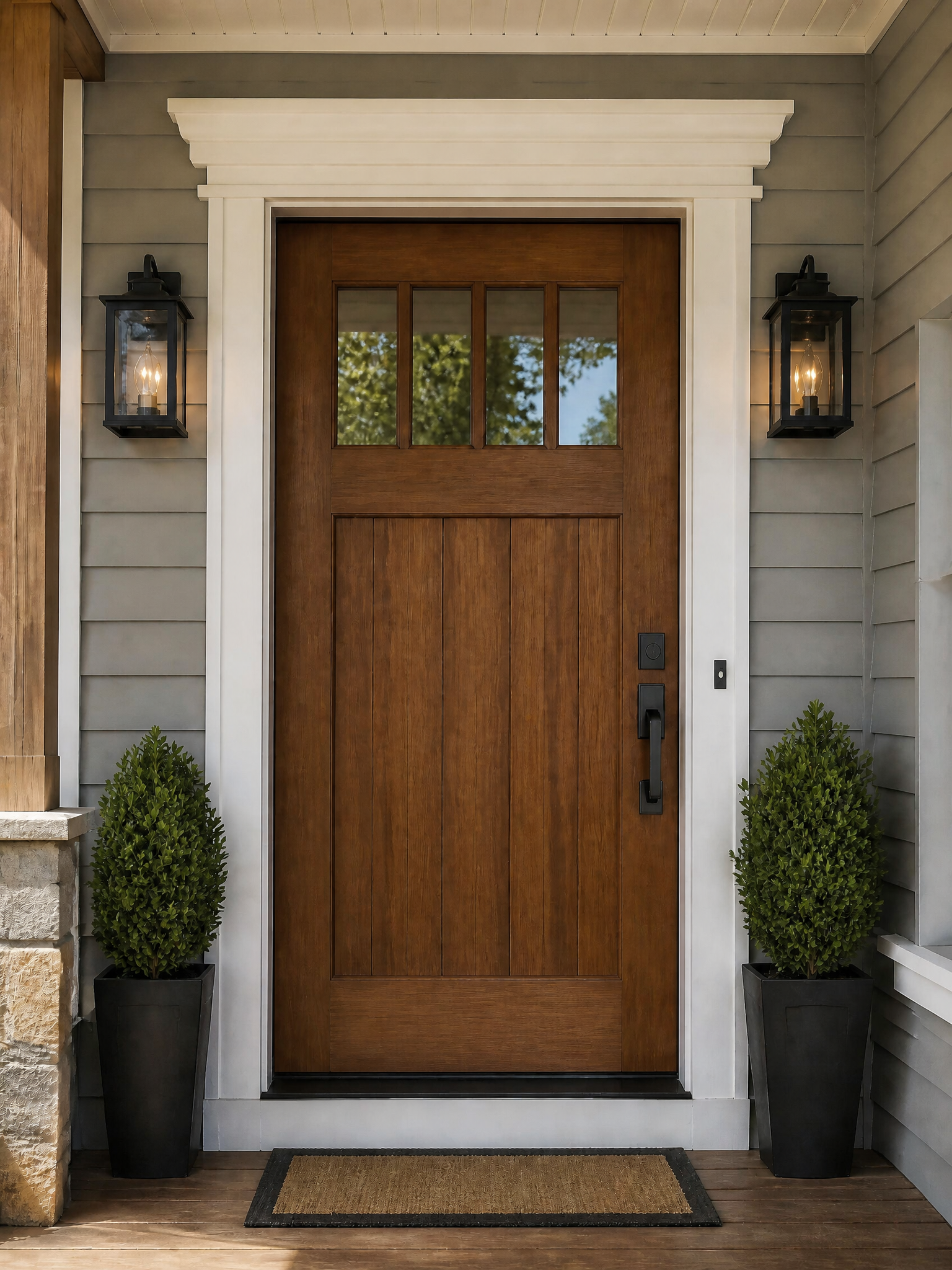A modern wooden front door with a six-pane top window, set in a white frame on a house porch with black lantern wall lights, potted topiary trees, and a woven doormat.