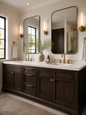 A double-sink bathroom vanity with dark wood cabinets, a white countertop, gold faucets and hardware, and two large rectangular mirrors. The room has warm beige walls, wall-mounted gold sconces, a window on the left, and a rug on the floor.